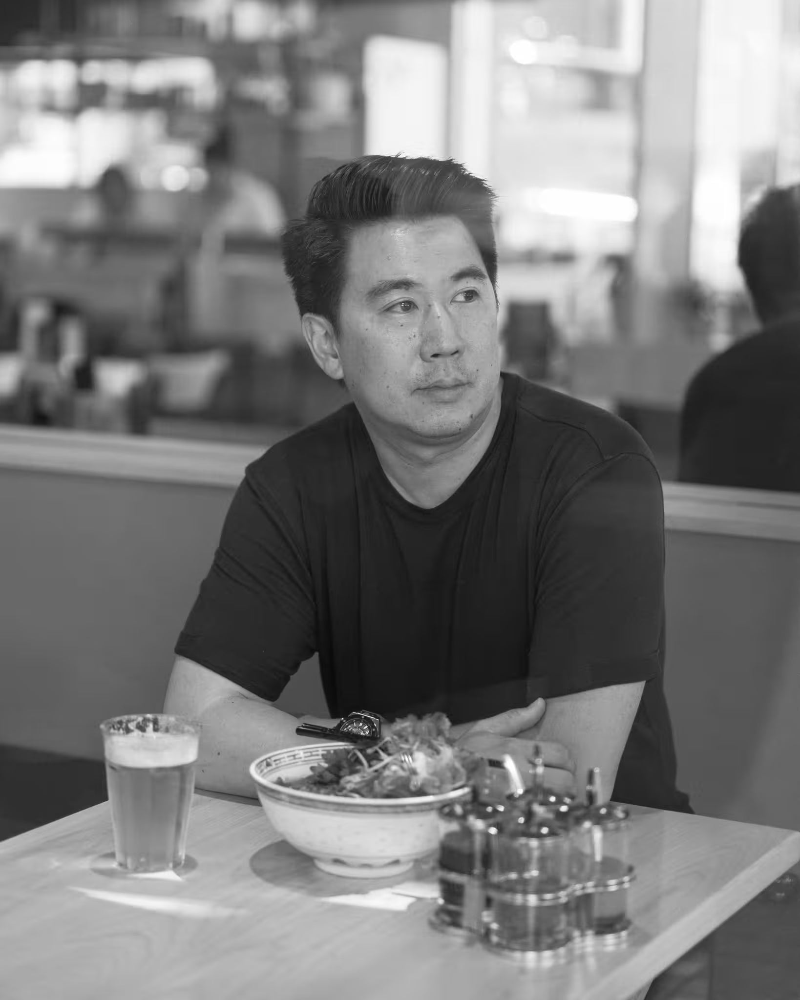 Black-and-white portrait of chef Angus An seated at a table in his restaurant with a bowl of noodles