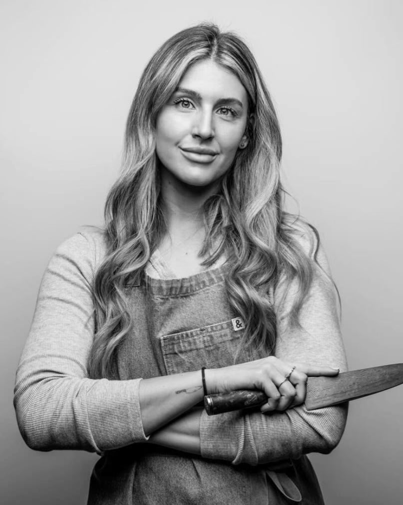Black-and-white portrait of chef Ashley Hoar in a denim apron, holding a chef's knife across her arms