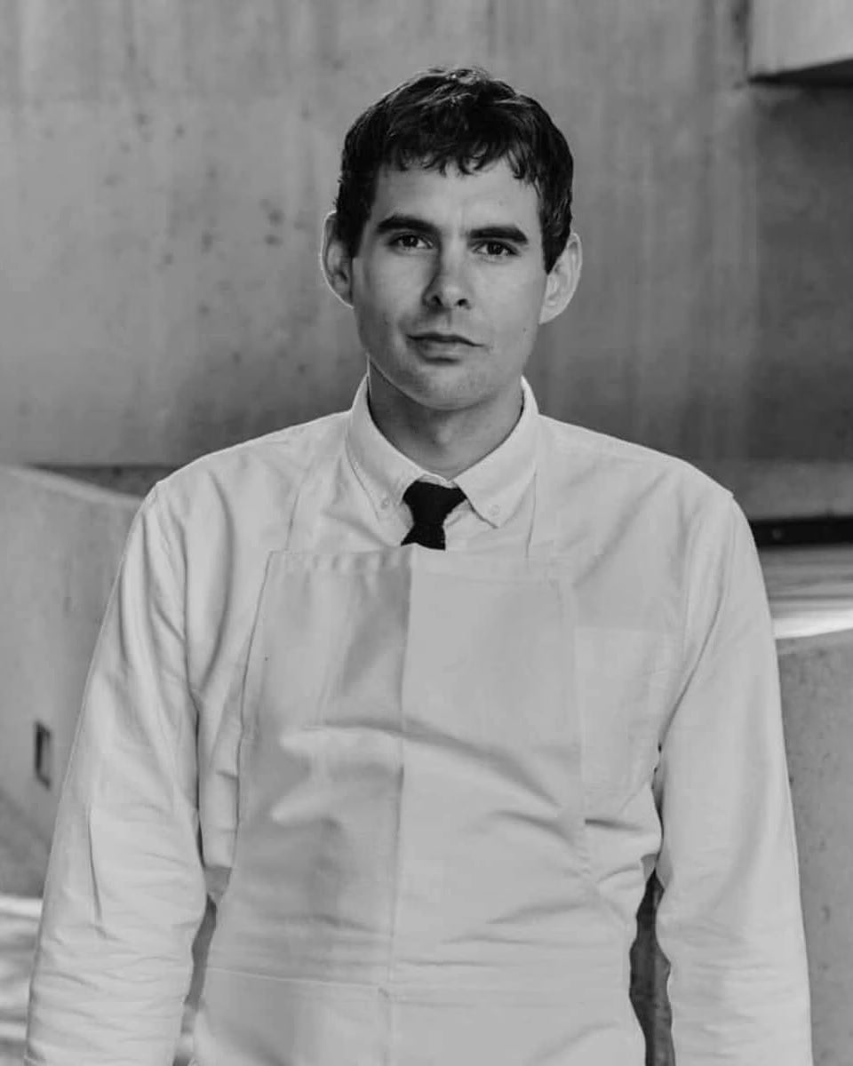 Black-and-white portrait of chef Francis Blais in a white shirt, tie, and apron against a concrete wall