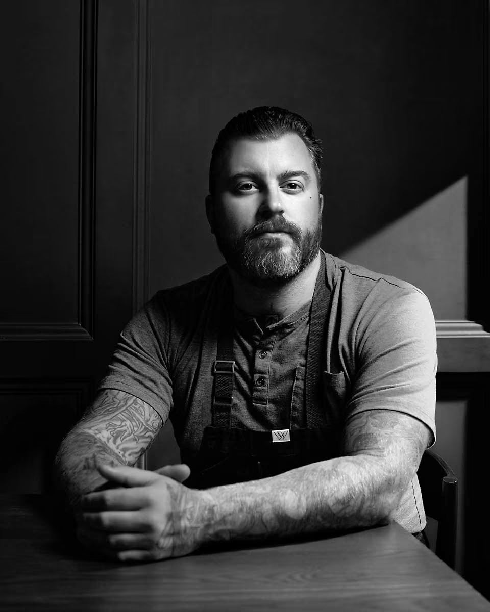 Black-and-white portrait of chef Gus Stieffenhofer-Brandson seated in an apron against a dark paneled wall