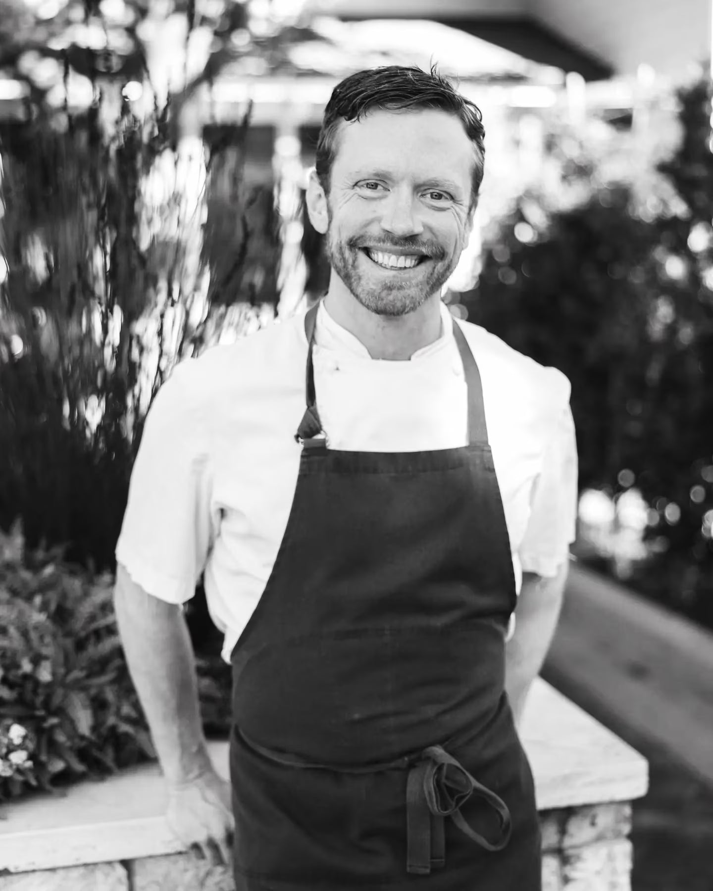 Black-and-white portrait of chef Jonny Black smiling, in chef's whites and a dark apron