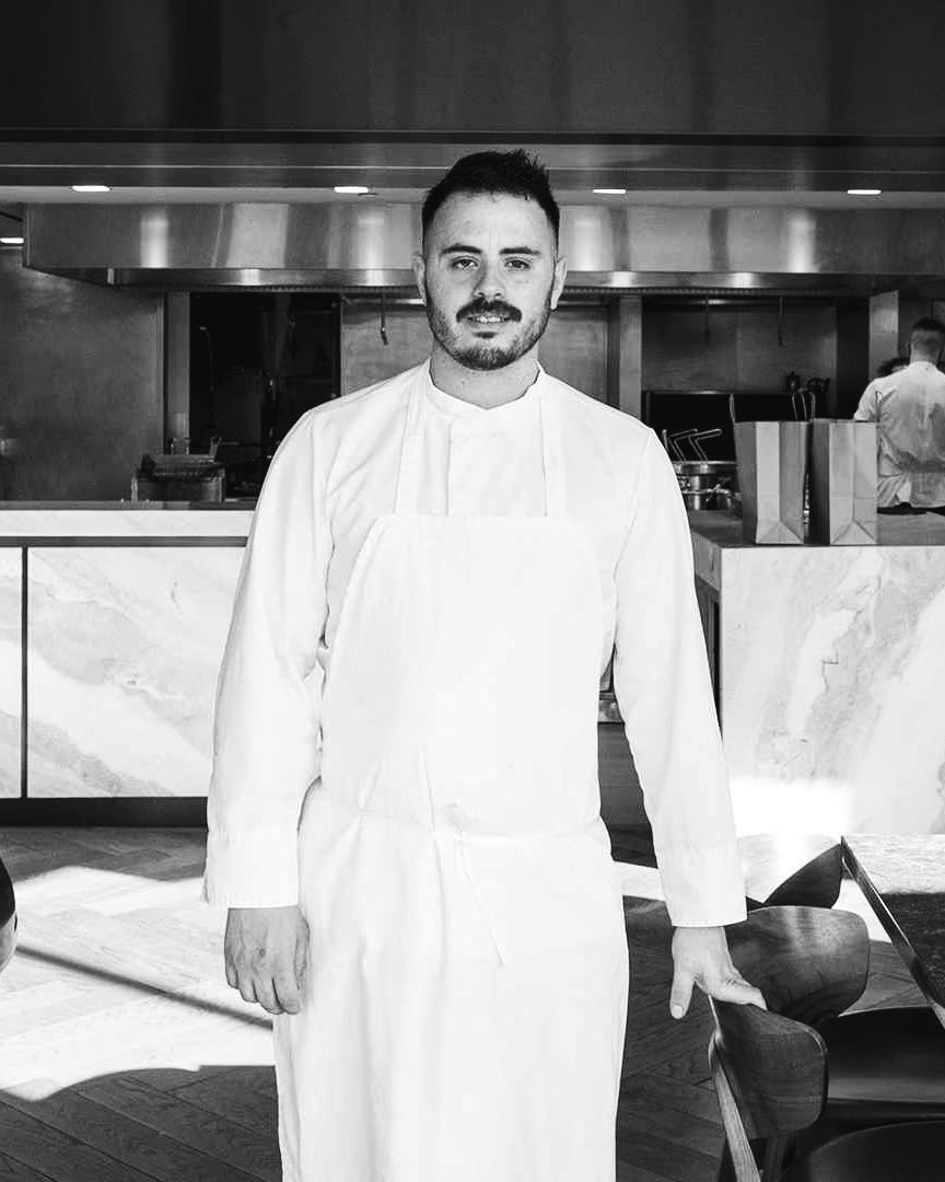 Black-and-white portrait of chef Rafa Covarrubias in chef's whites in a fine-dining open kitchen