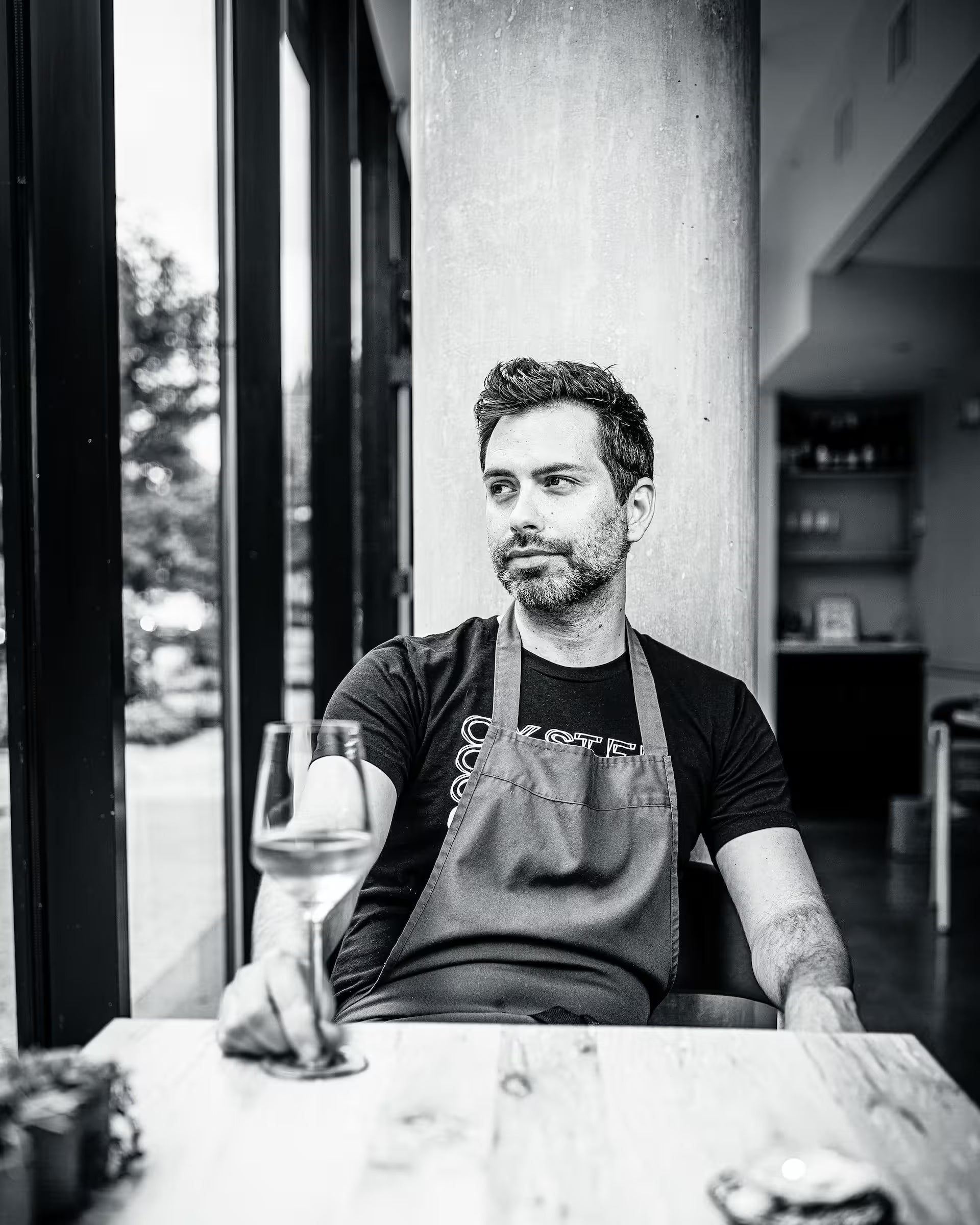 Black-and-white portrait of chef Rob Rubba seated at a restaurant table by large windows, with a glass of wine