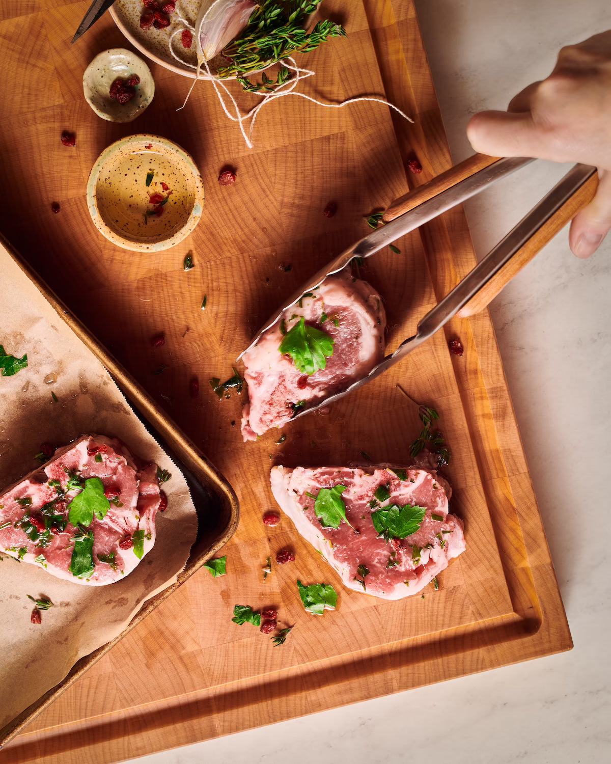 Hand grabbing meat chops with tongs on maple cutting board with herbs and baking tray