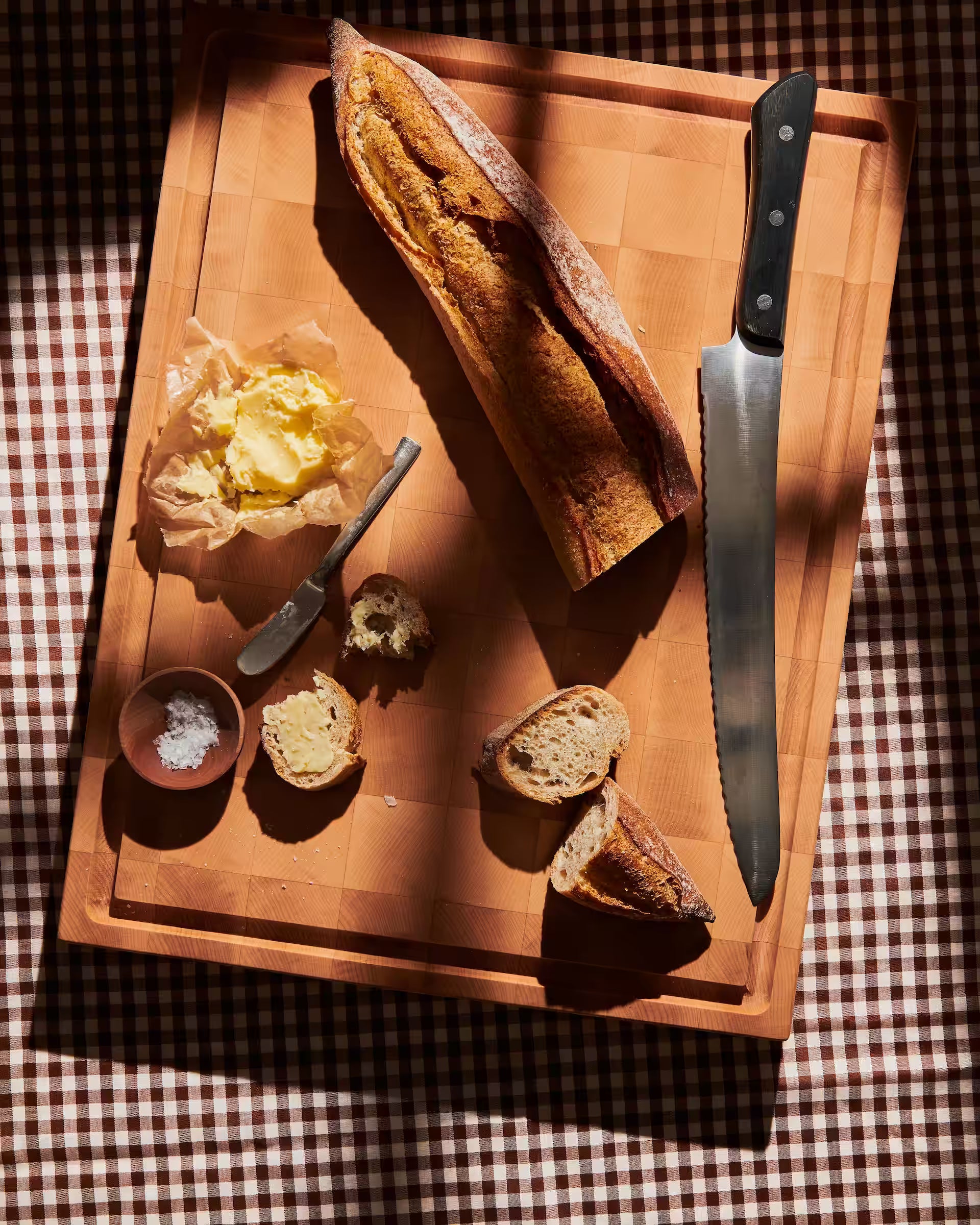Maple end-grain cutting board with a sliced baguette and butter on parchment, sunlit on a gingham cloth
