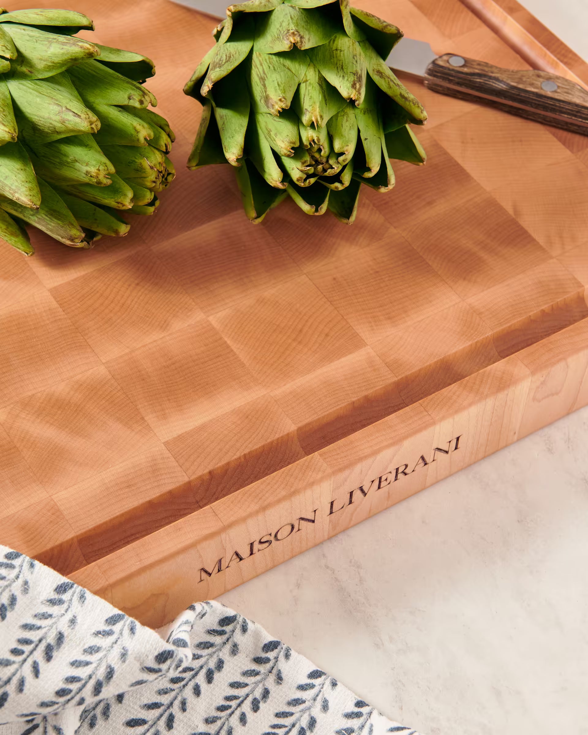 Maison Liverani name engraved on the side of a maple end-grain cutting board beside two artichokes