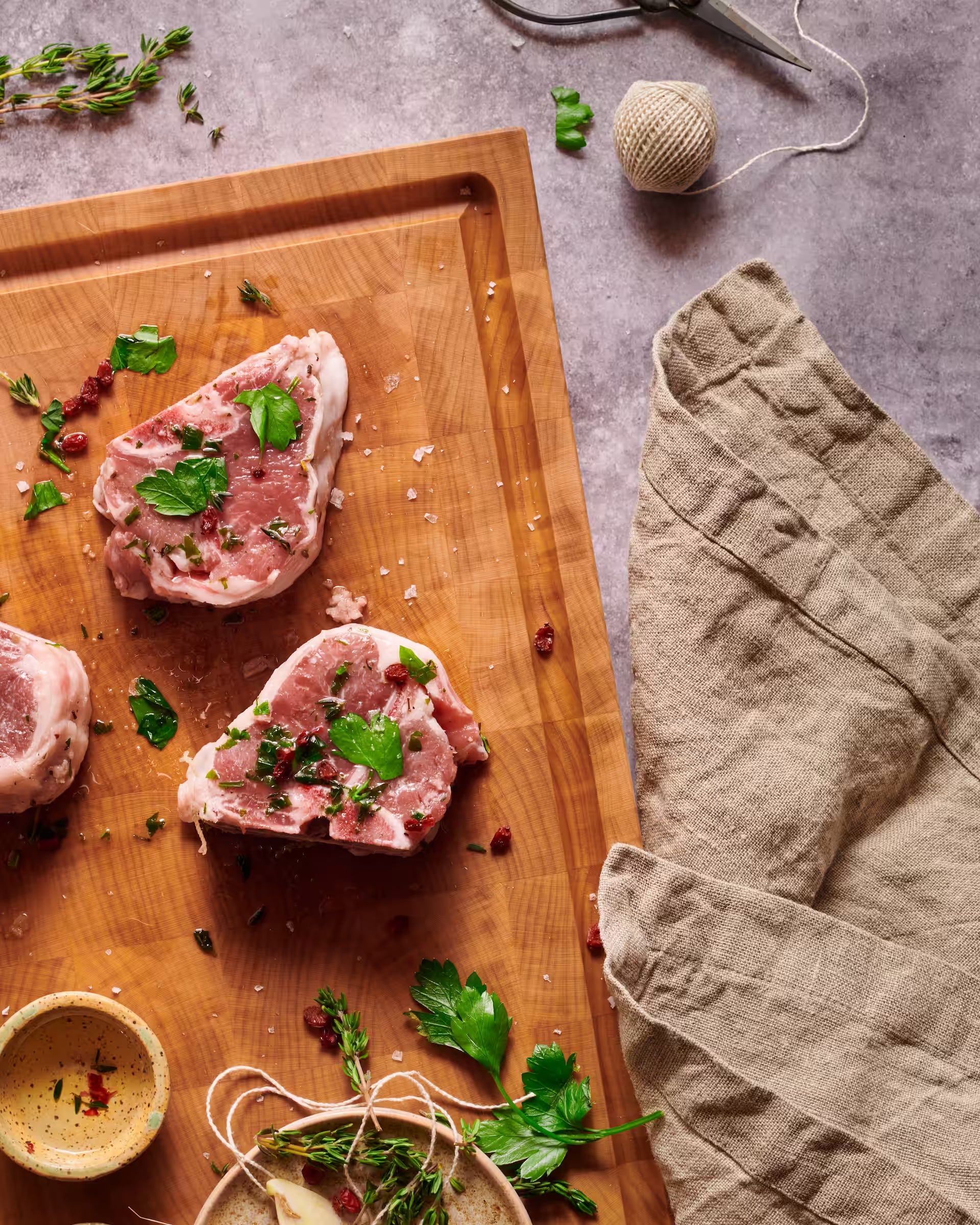 Maple end-grain cutting board with two raw pork chops seasoned with parsley and pink peppercorns
