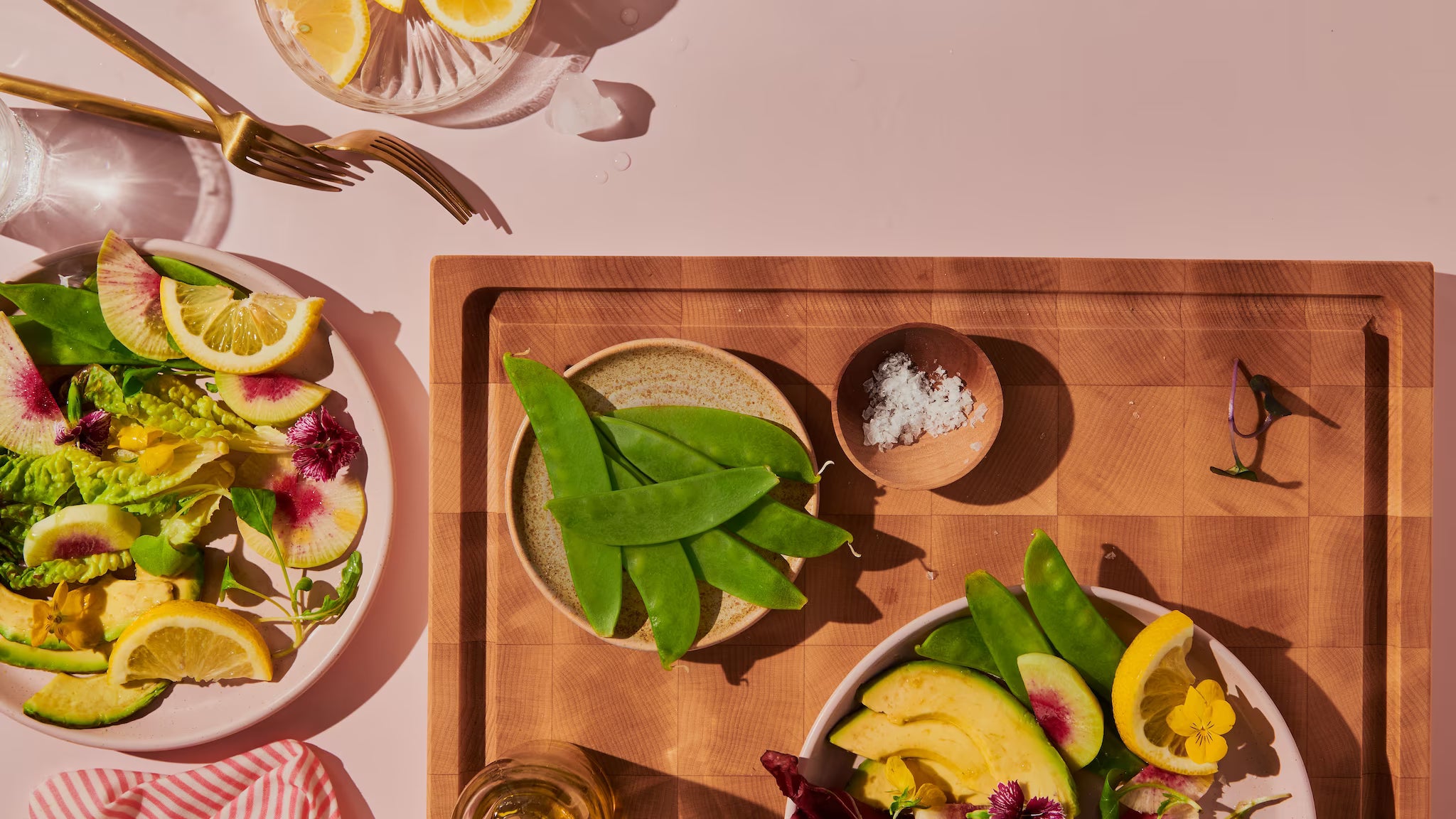 Maple end-grain cutting board with snow peas and flaky salt beside a fresh salad in sunlight