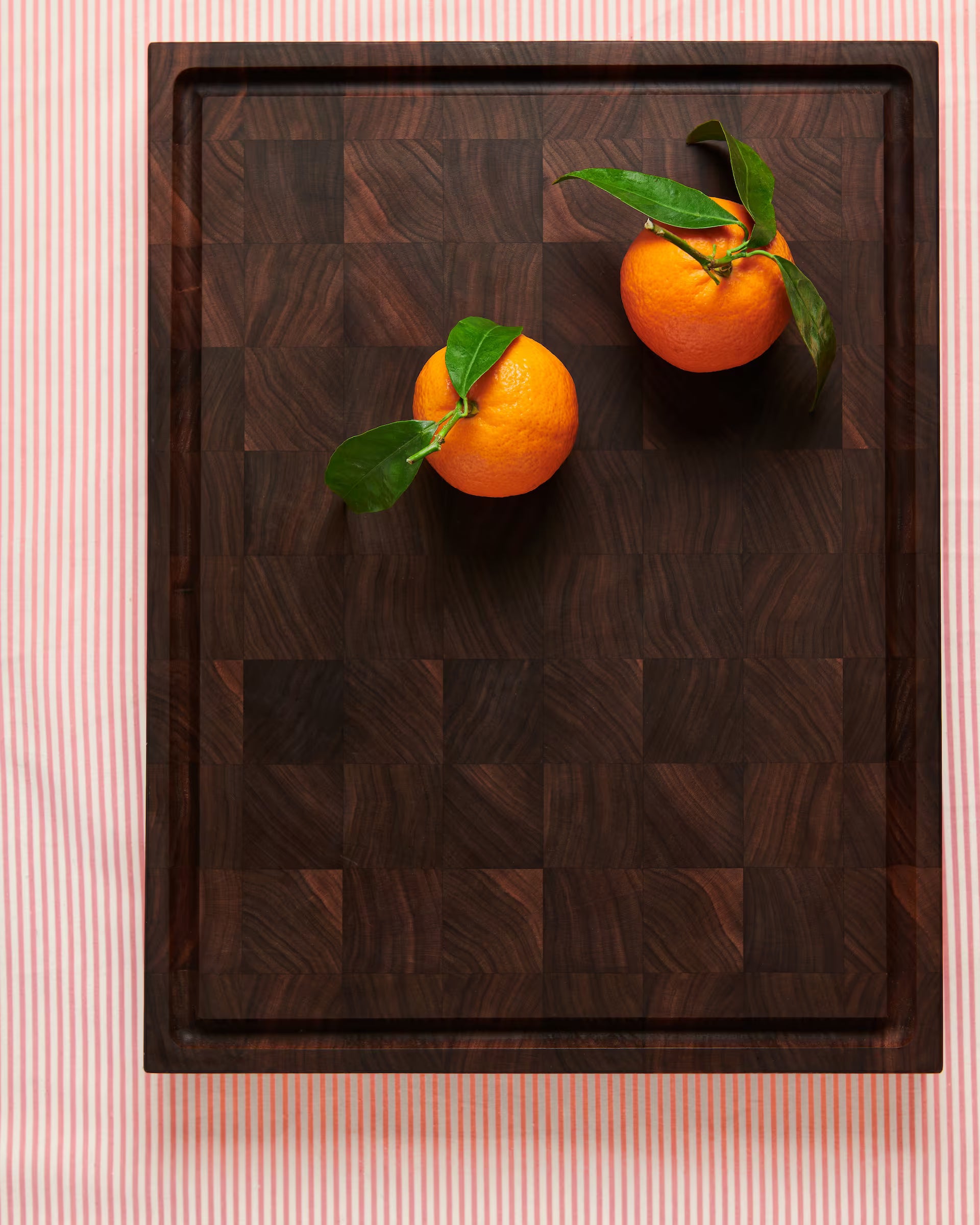 Maison Liverani walnut end-grain cutting board with two leafy whole oranges on a striped cloth